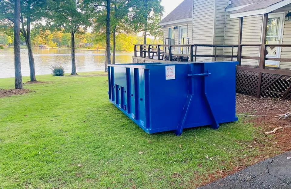Blue roll-off dumpster set on grass beside a lakeside deck