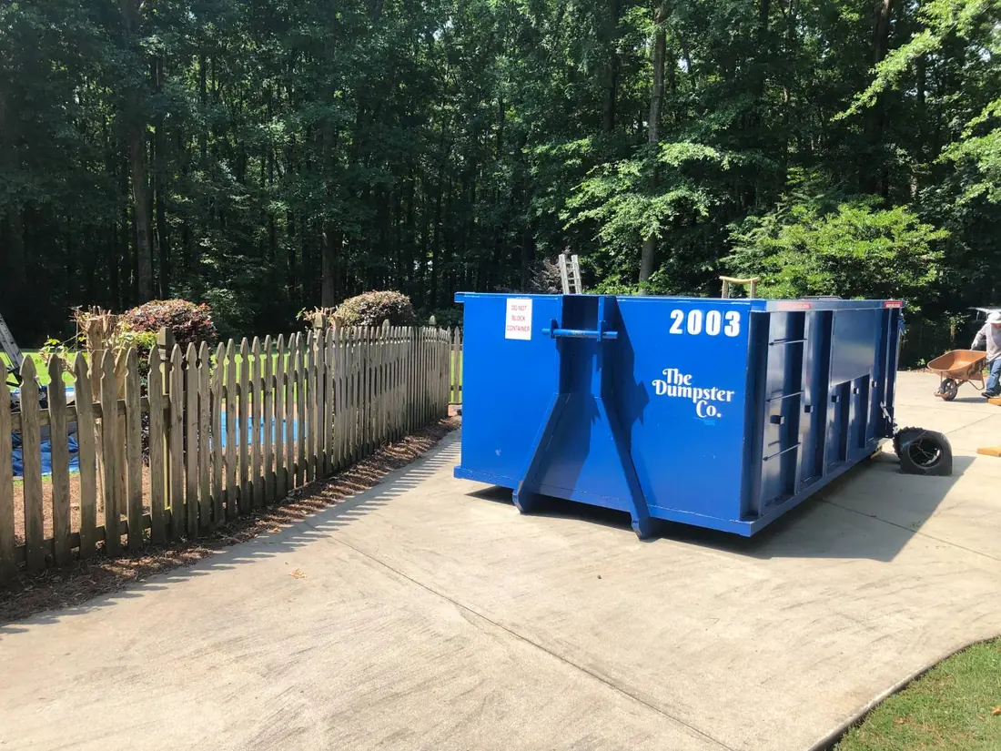 The Dumpster Co. roll-off dumpster on a concrete driveway beside a backyard fence