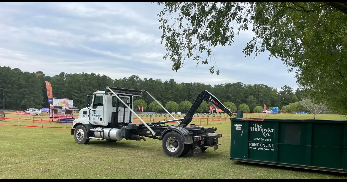 Roll-off truck delivering a green The Dumpster Co. dumpster to an outdoor event field