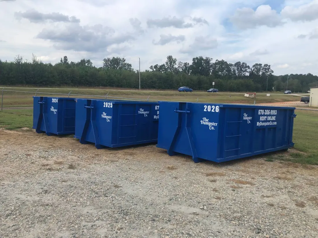 Three blue The Dumpster Co. roll-off dumpsters staged on a gravel lot